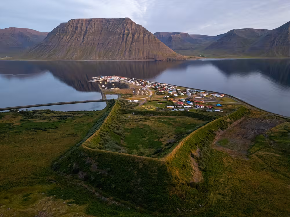 Aerial view of small Westfjords village on a narrow peninsula surrounded by calm fjord waters and dramatic flat-topped mountains