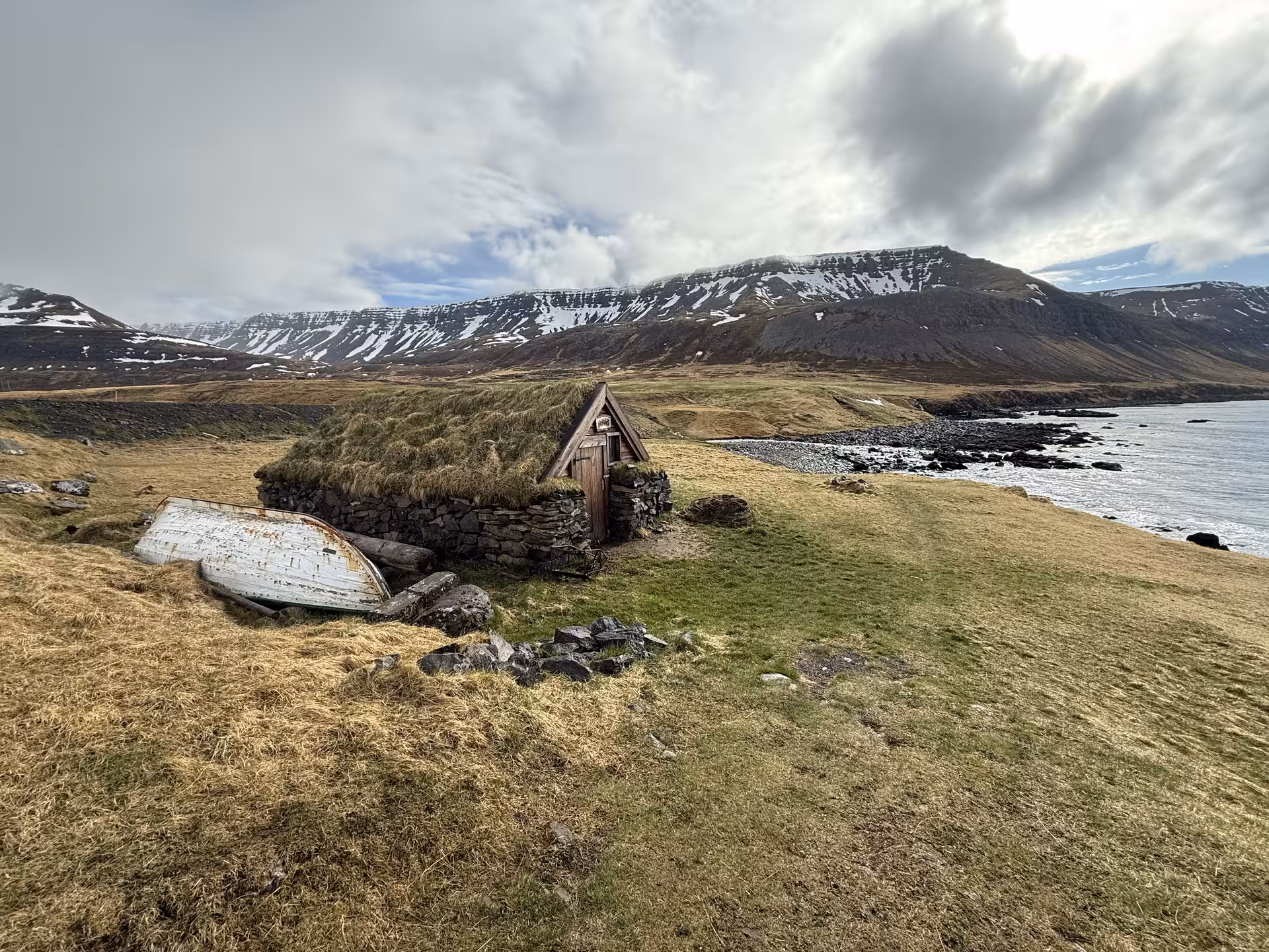 Traditional Icelandic turf house with stone walls and grass roof near fjord coastline, snow-capped Westfjords mountains behind
