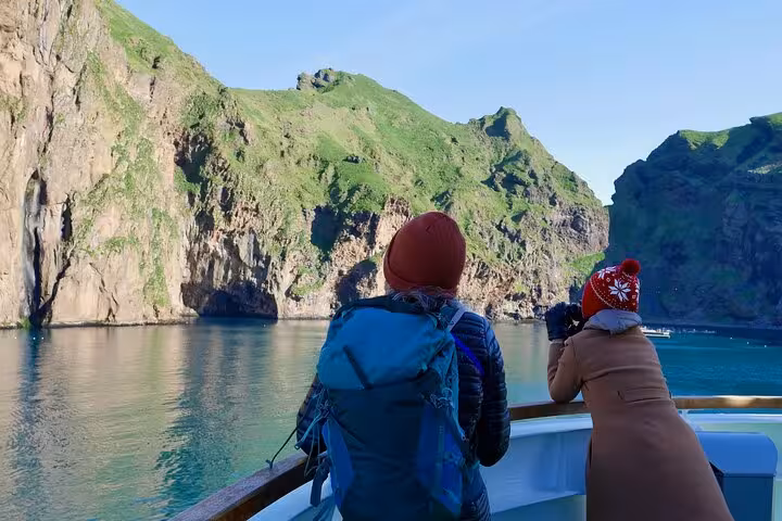 Two travelers on a boat viewing dramatic green cliffs and sea caves of Westman Islands, Iceland