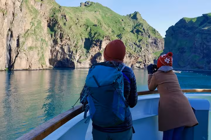 Two tourists on a boat deck viewing dramatic green cliffs and turquoise waters of Westman Islands