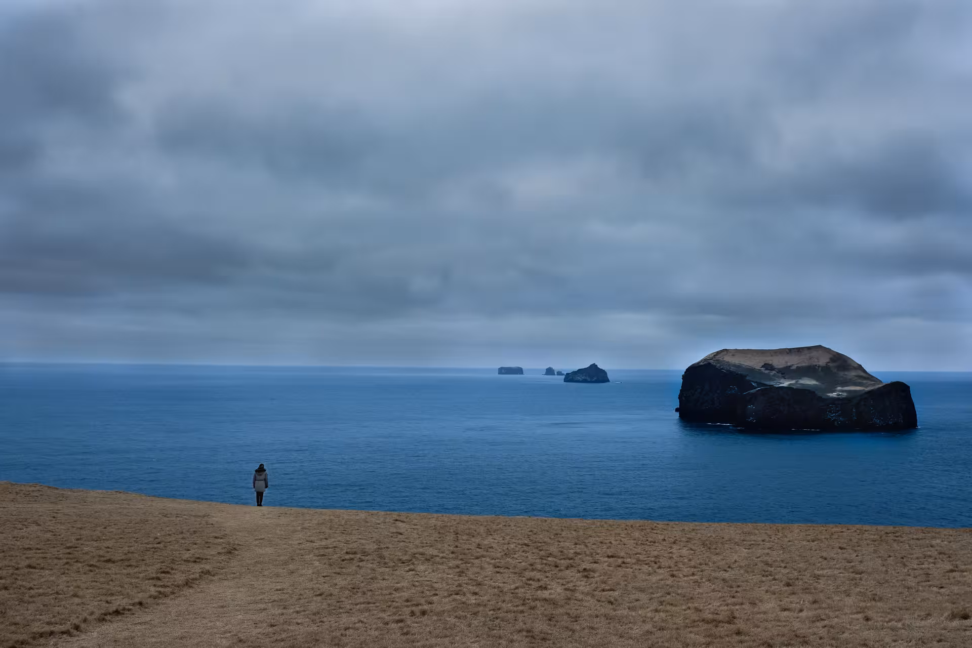 Lone traveler standing on grassy cliffs overlooking Westman Islands volcanic rock formations in blue ocean