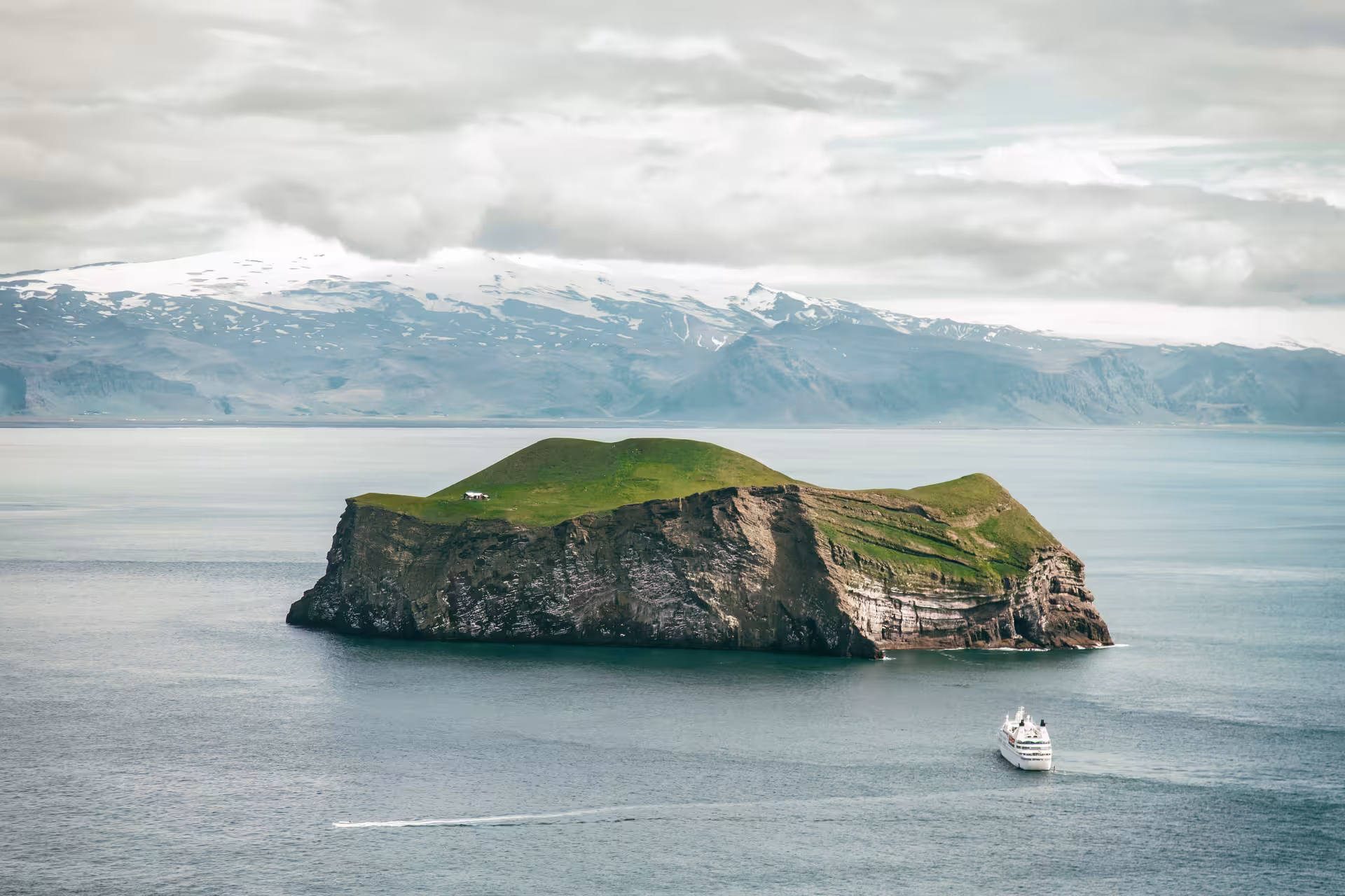 Cruise ship passing Elliðaey island in the Westman Islands with snow-capped Icelandic mountains behind