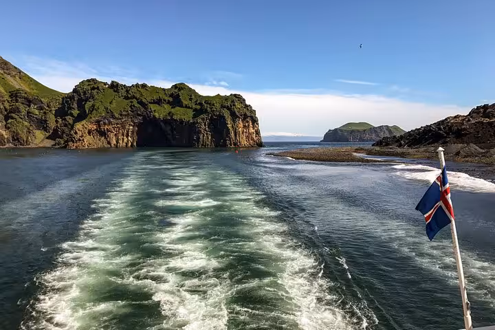 Boat wake leaving Westman Islands rocky cliffs with Icelandic flag, heading to Reykjavik