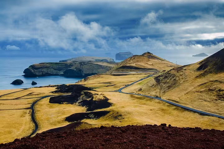 Scenic winding road through golden volcanic landscape of Westman Islands with ocean views and dramatic clouds