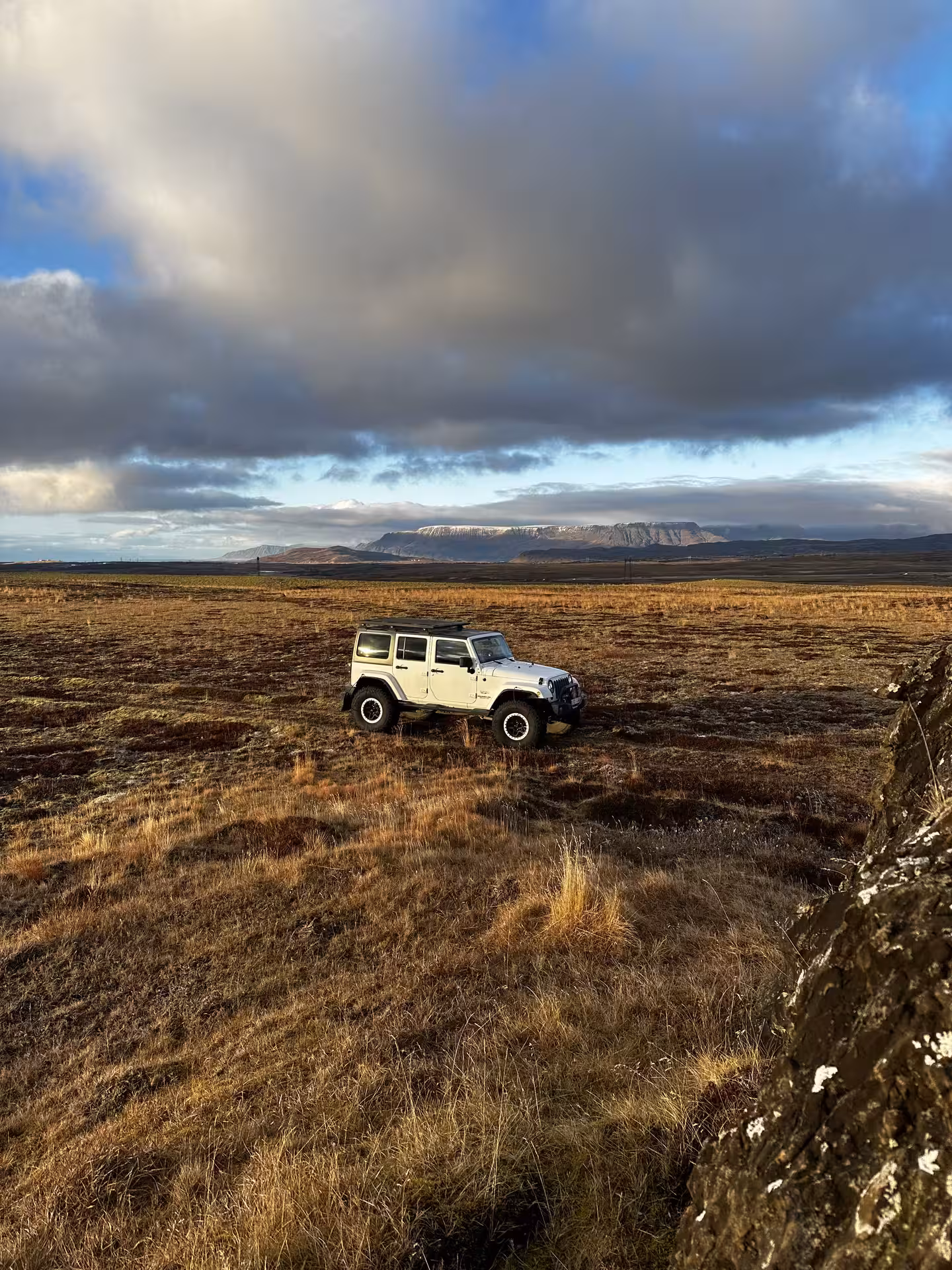 White Jeep Wrangler on Icelandic highland tundra with dramatic clouds and snow-capped mountains in background