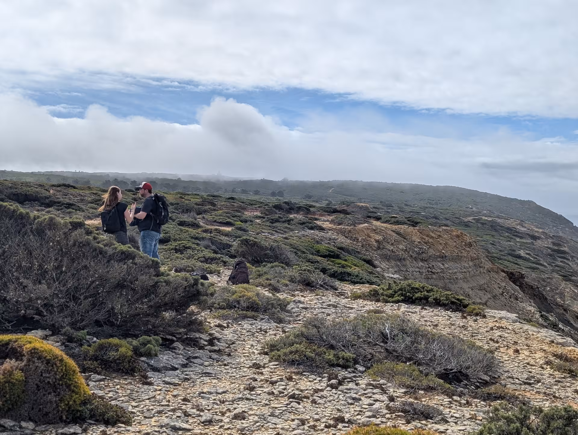 Couple with backpacks on rugged Wild West Coast headland trail, foggy sky and ocean views on guided hike