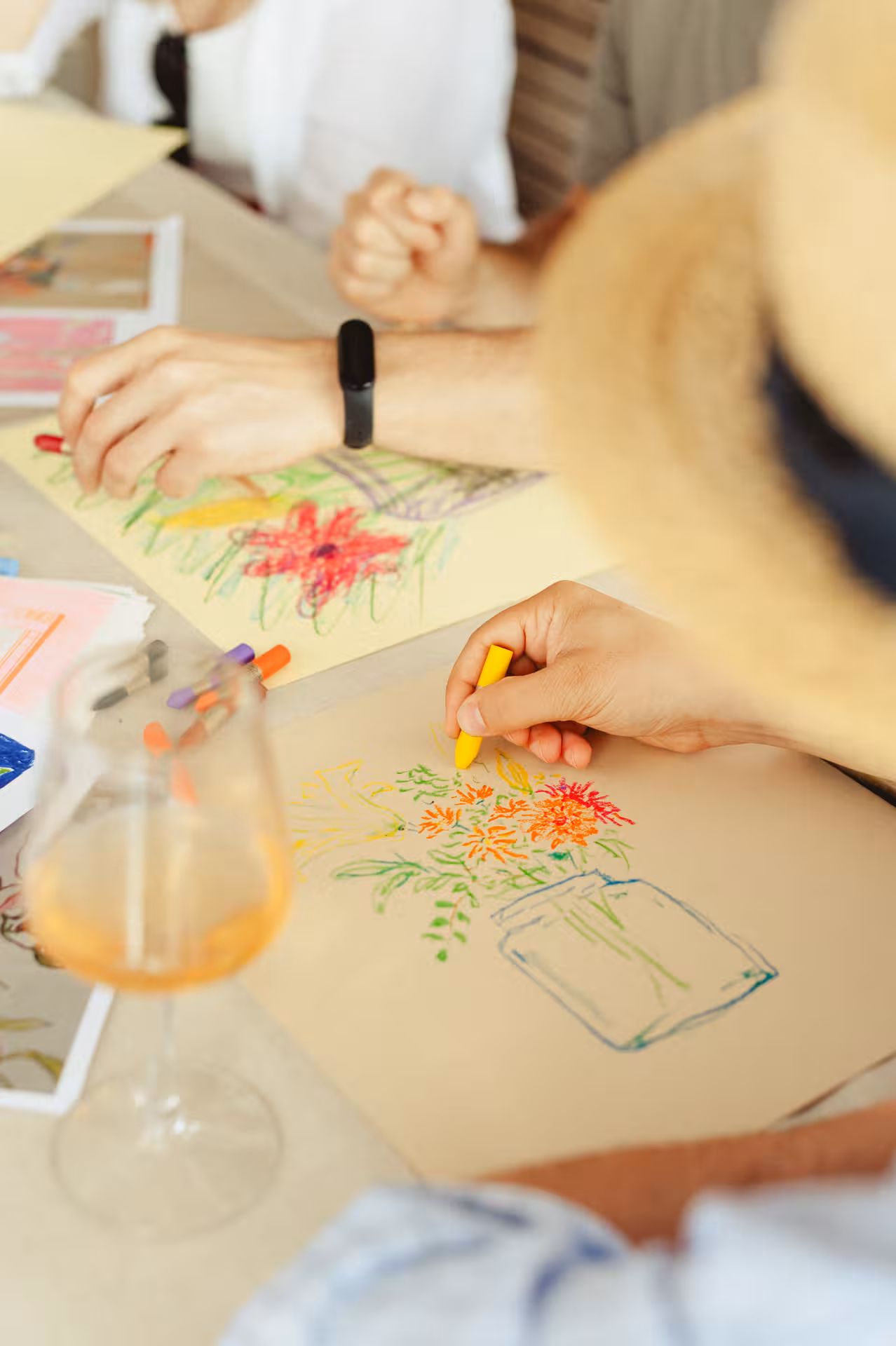 Close-up of hands sketching colorful flowers with pastels beside a wine glass at a Wine & Art workshop