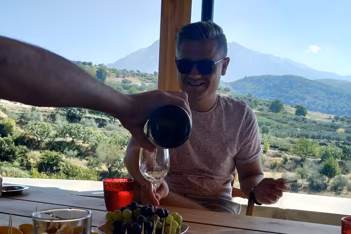 A man smiling as wine is poured into his glass with scenic Berat vineyards in the background.