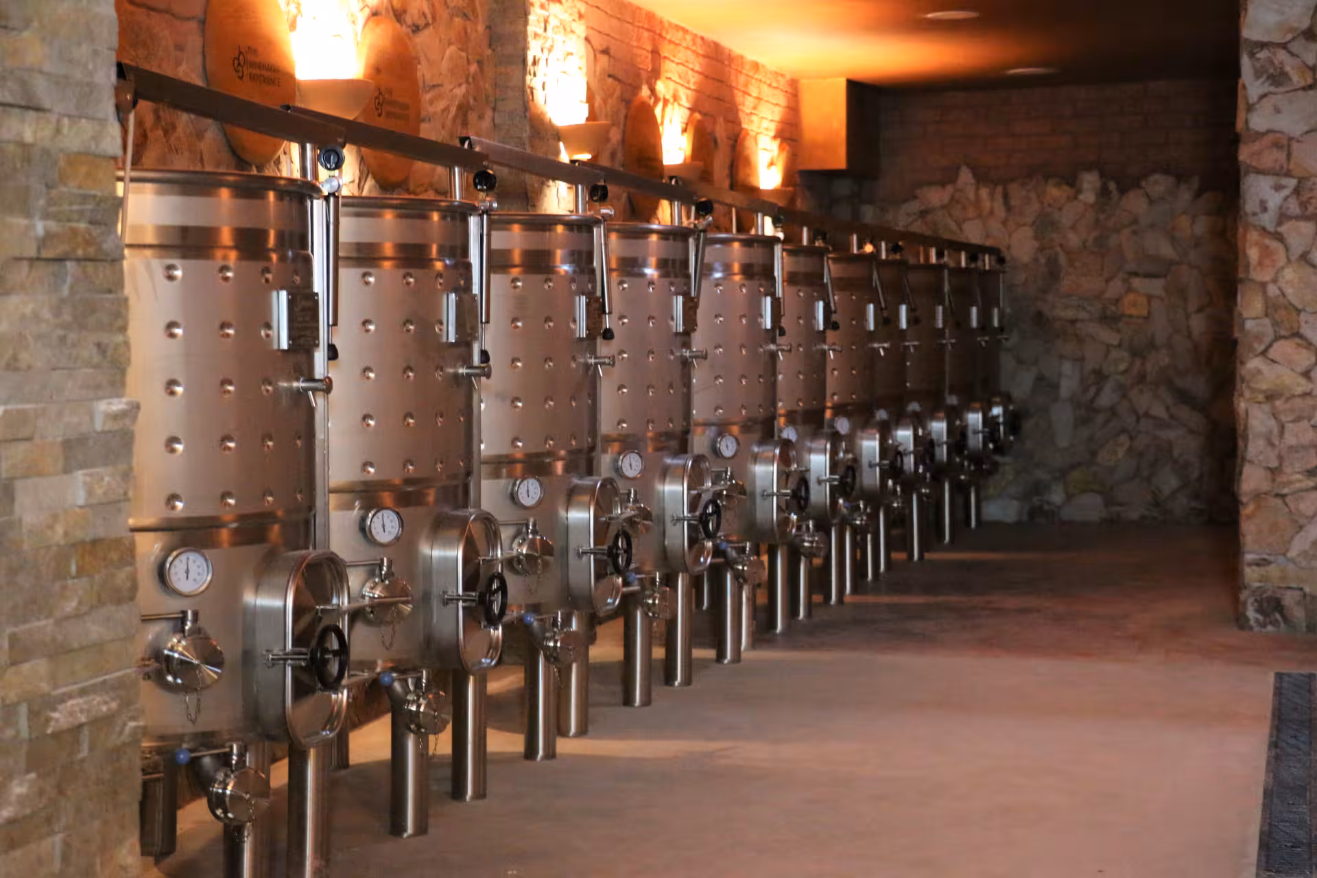 Row of stainless steel wine fermentation tanks in a rustic stone cellar on a guided winery tour