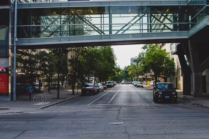 Glass skybridge over quiet Winnipeg downtown street with parked cars and green trees