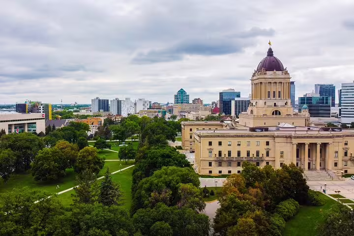 Aerial view of Manitoba Legislative Building with green parklands and Winnipeg skyline under cloudy skies