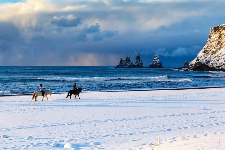 Two riders on horseback crossing a snow-covered black sand beach with dramatic sea stacks in Iceland
