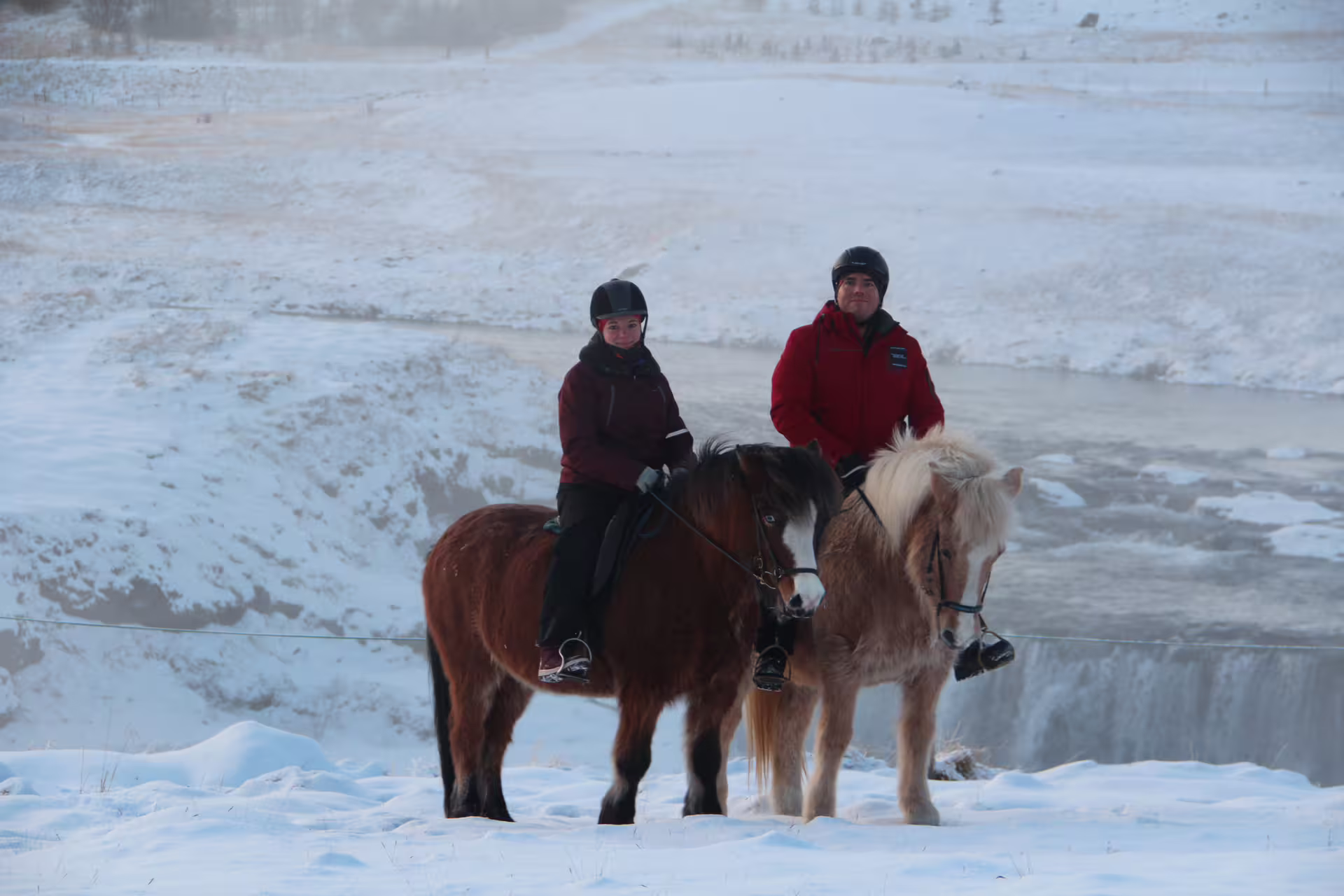 Two riders on Icelandic horses in snowy Skagafjordur landscape with frozen waterfall in background