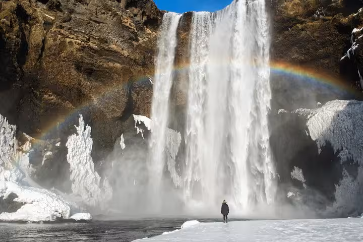 Person standing before massive Skógafoss waterfall with rainbow and snow-covered ground in winter Iceland