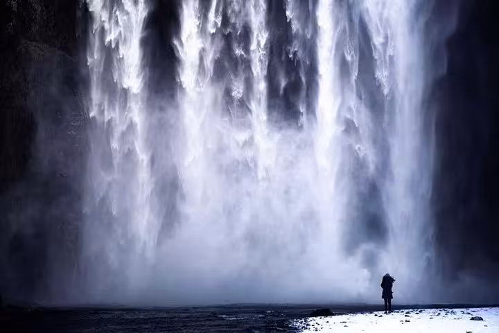 Lone figure standing before massive Skógafoss waterfall in winter, surrounded by mist and snow