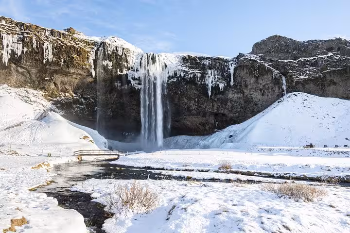Seljalandsfoss waterfall cascading over snow-covered cliffs in winter with icicles and a wooden bridge below