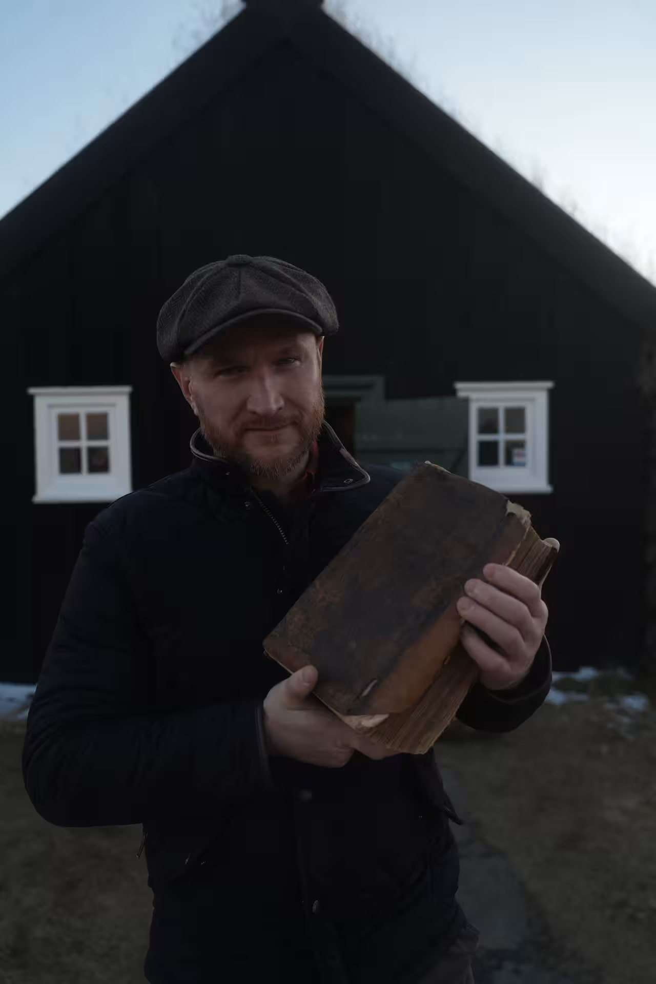 Iceland witchcraft tour guide holding ancient spell book outside traditional black Icelandic turf house in winter