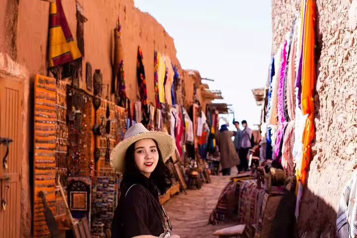 Woman exploring Ait Benhaddou souk alley with colorful textiles on Morocco 9 days tour from Marrakech itinerary