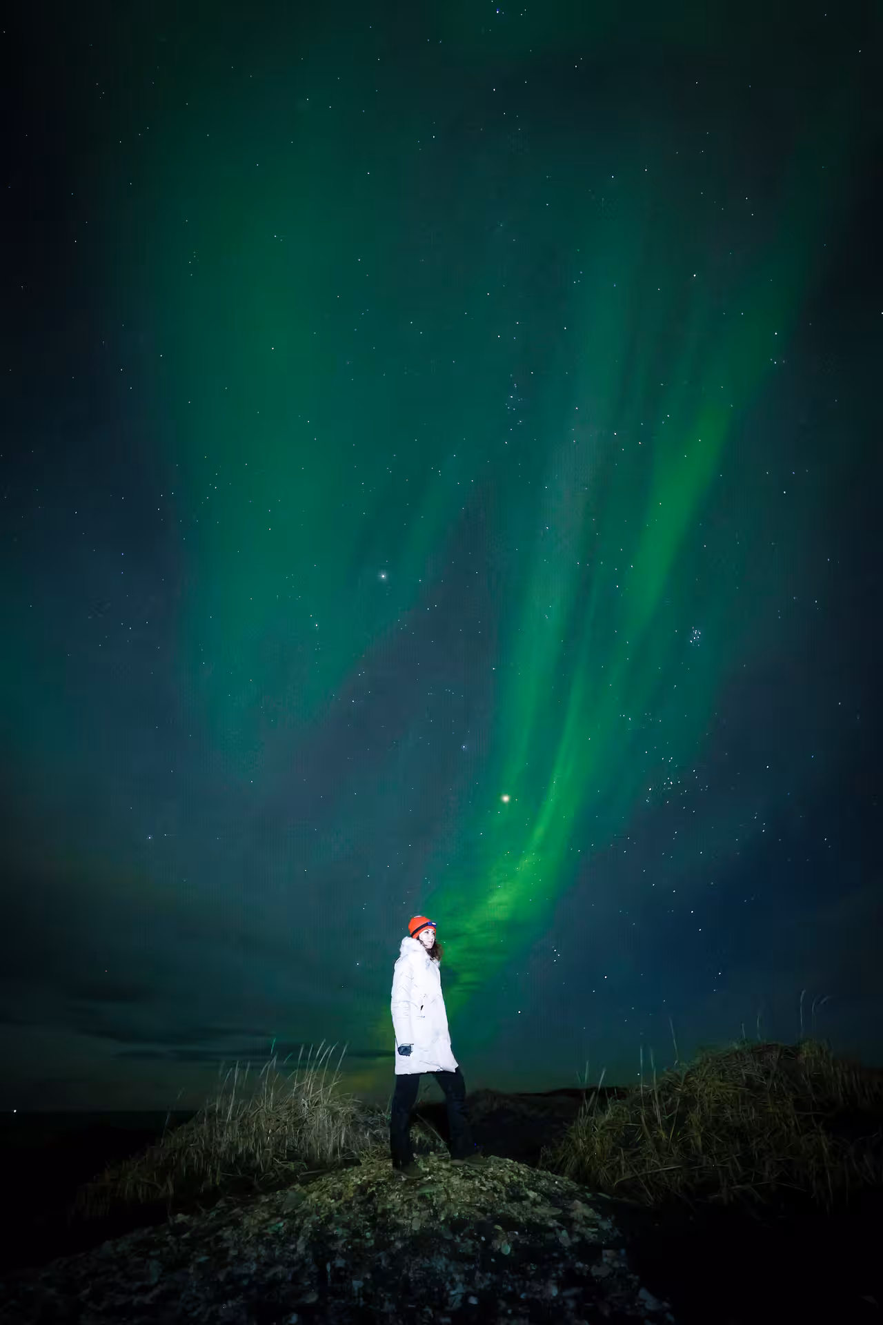Woman in white winter coat stands on rocky hill beneath vivid green aurora borealis northern lights night sky