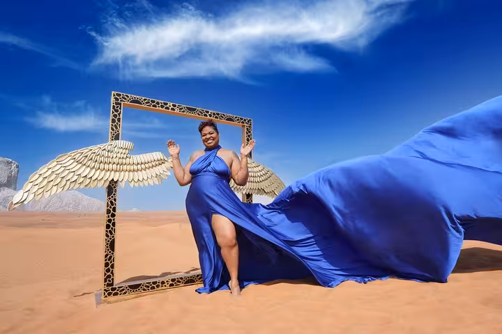 Woman in a flowing blue dress poses with angel wings in the expansive Dubai desert under a clear sky.