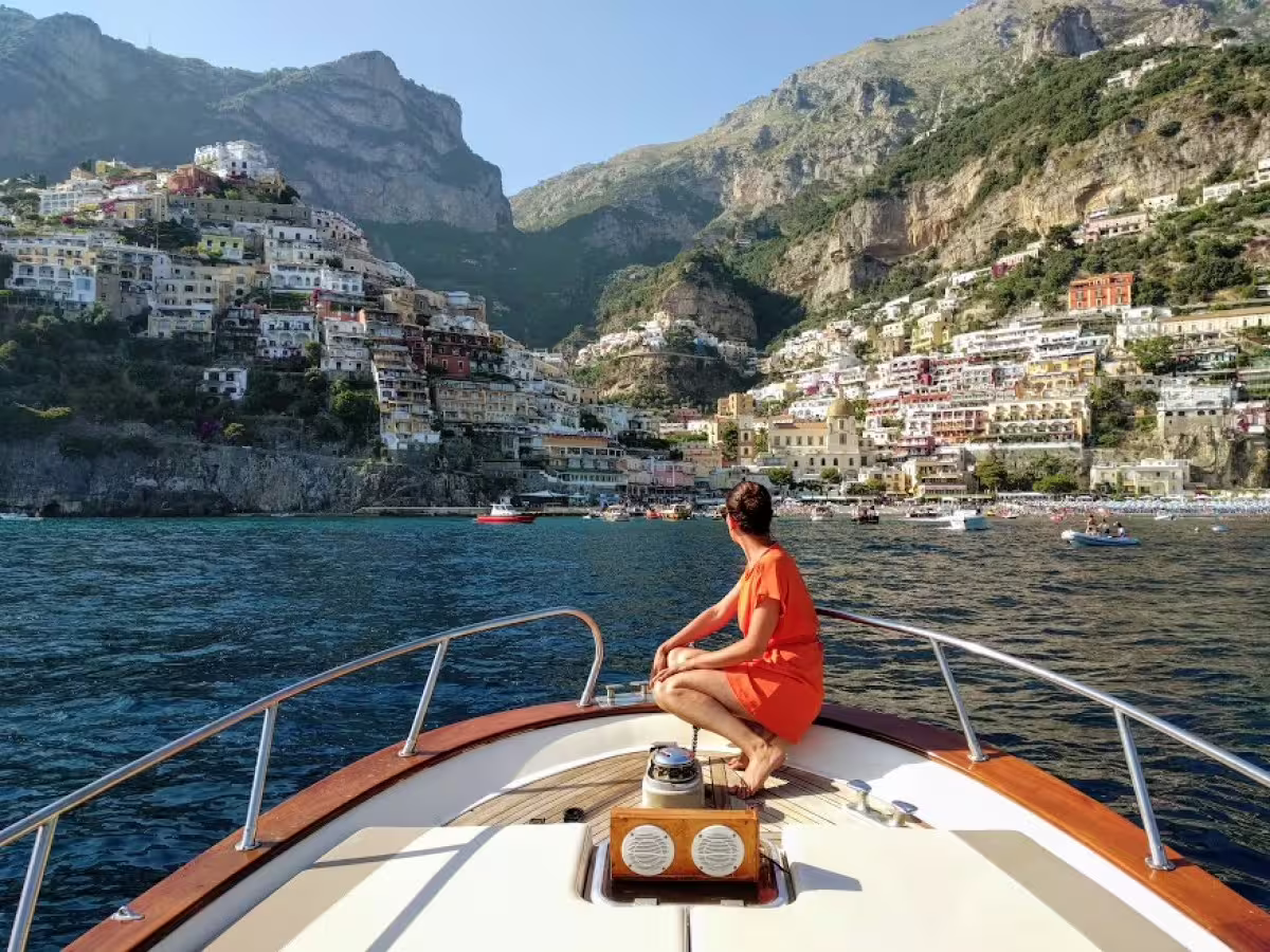Woman in orange dress relaxing on boat bow approaching colorful Positano cliffs on Sorrento and Amalfi Coast tour