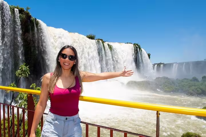 Smiling woman poses on a viewing platform with a stunning view of the Brazilian Falls on a clear sunny day.