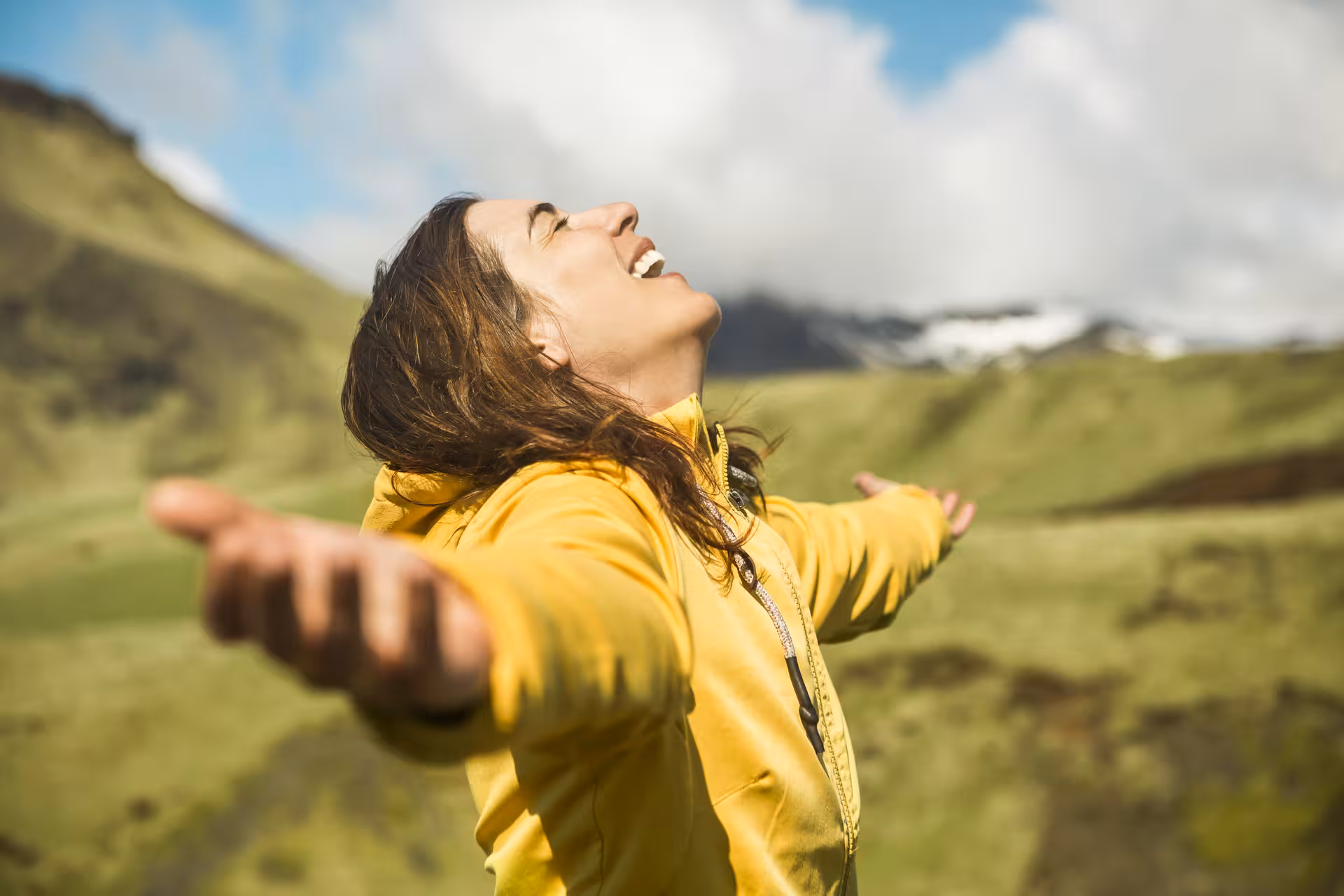 Woman in yellow jacket with arms outstretched, laughing in Iceland's green mountain landscape