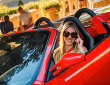 Smiling woman in sunglasses driving a red Ferrari convertible supercar on a sunny day