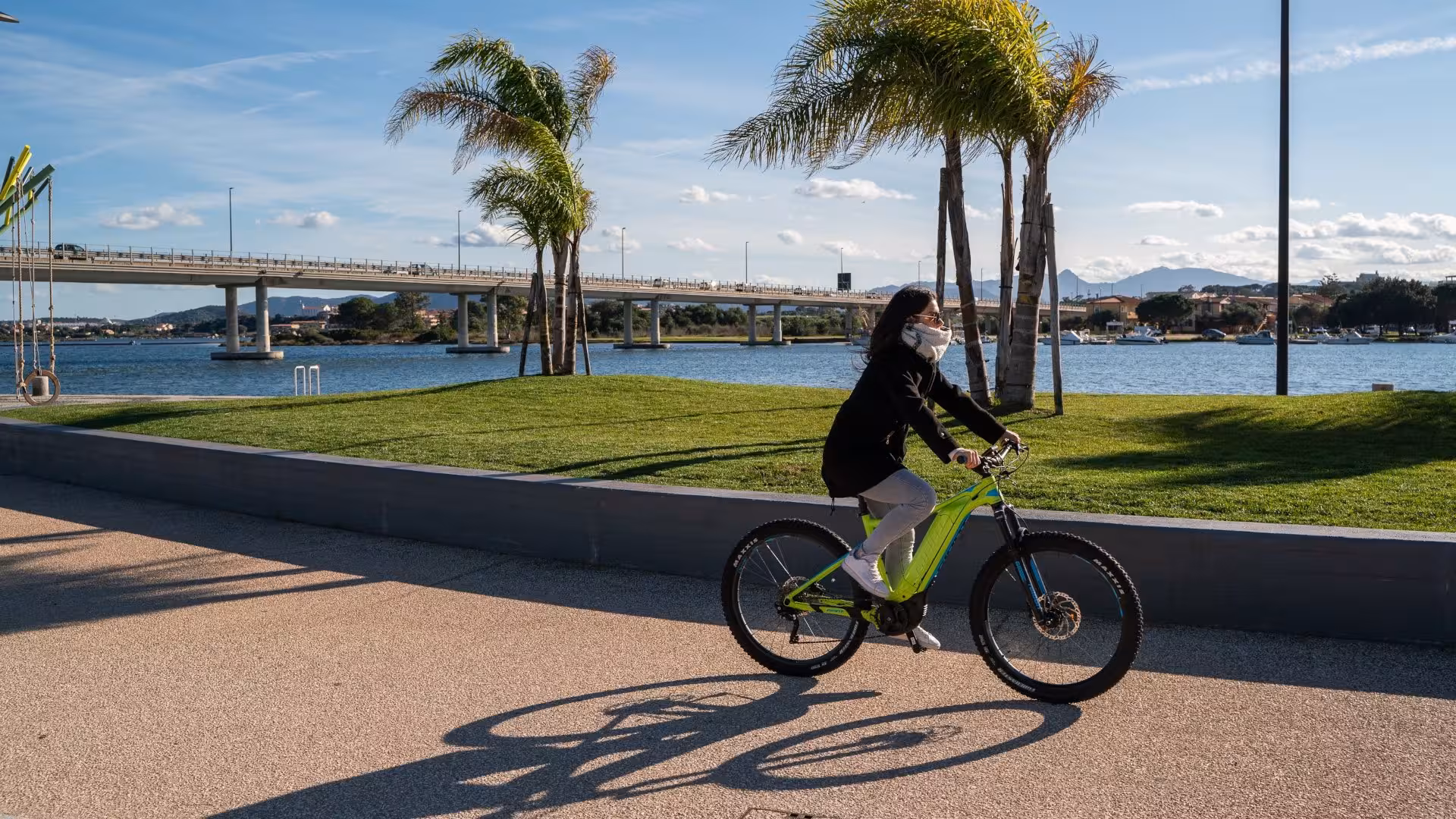 Woman riding an e-bike along the waterfront in Olbia, Sardinia, with a scenic bridge and palm trees in the background.