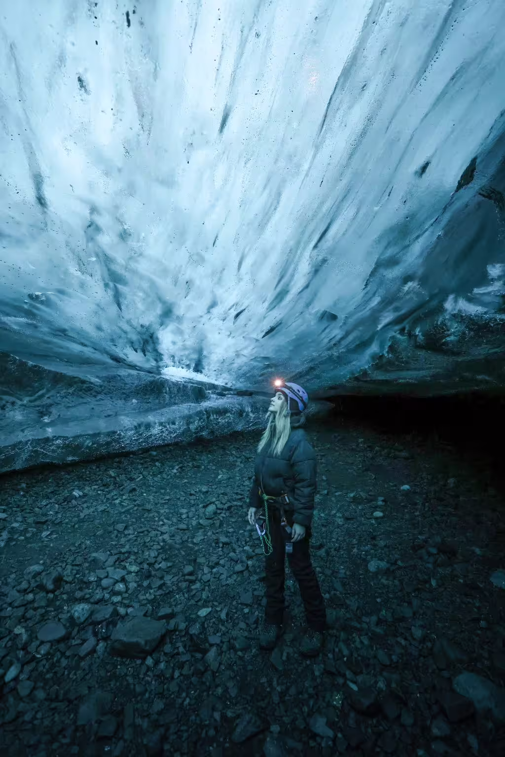 Woman with headlamp and climbing gear gazing up at massive blue glacial ice walls on Iceland ice cave guided tour