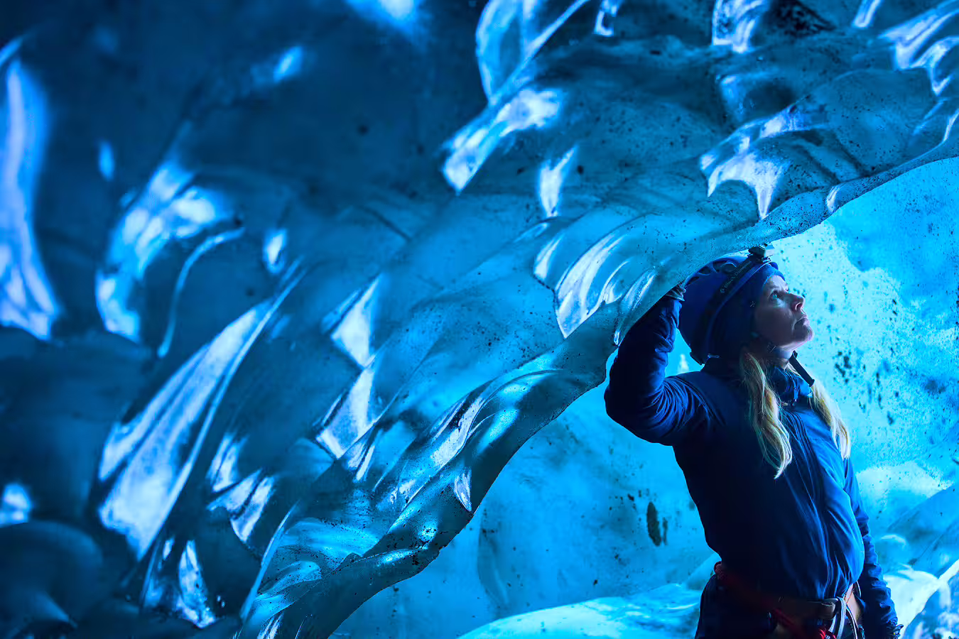 Woman in blue jacket touching glacial ice cave walls glowing in vivid blue light during guided tour