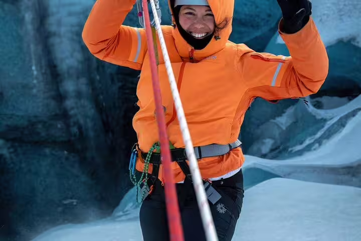 Joyful woman in orange jacket and climbing harness exploring glacier caves on Iceland adventure tour
