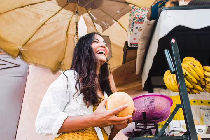 Smiling woman holding fruit at a lively market stall under an umbrella in Marrakesh, Egypt.