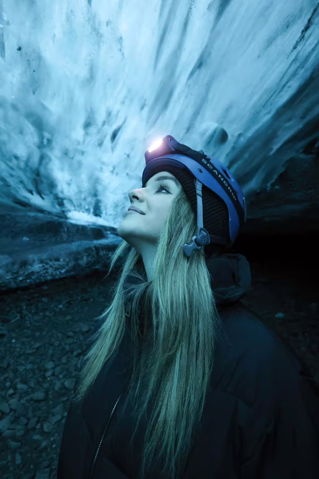 Woman with helmet and headlamp gazes in awe at glowing blue ice cave walls on a private early bird tour in Iceland
