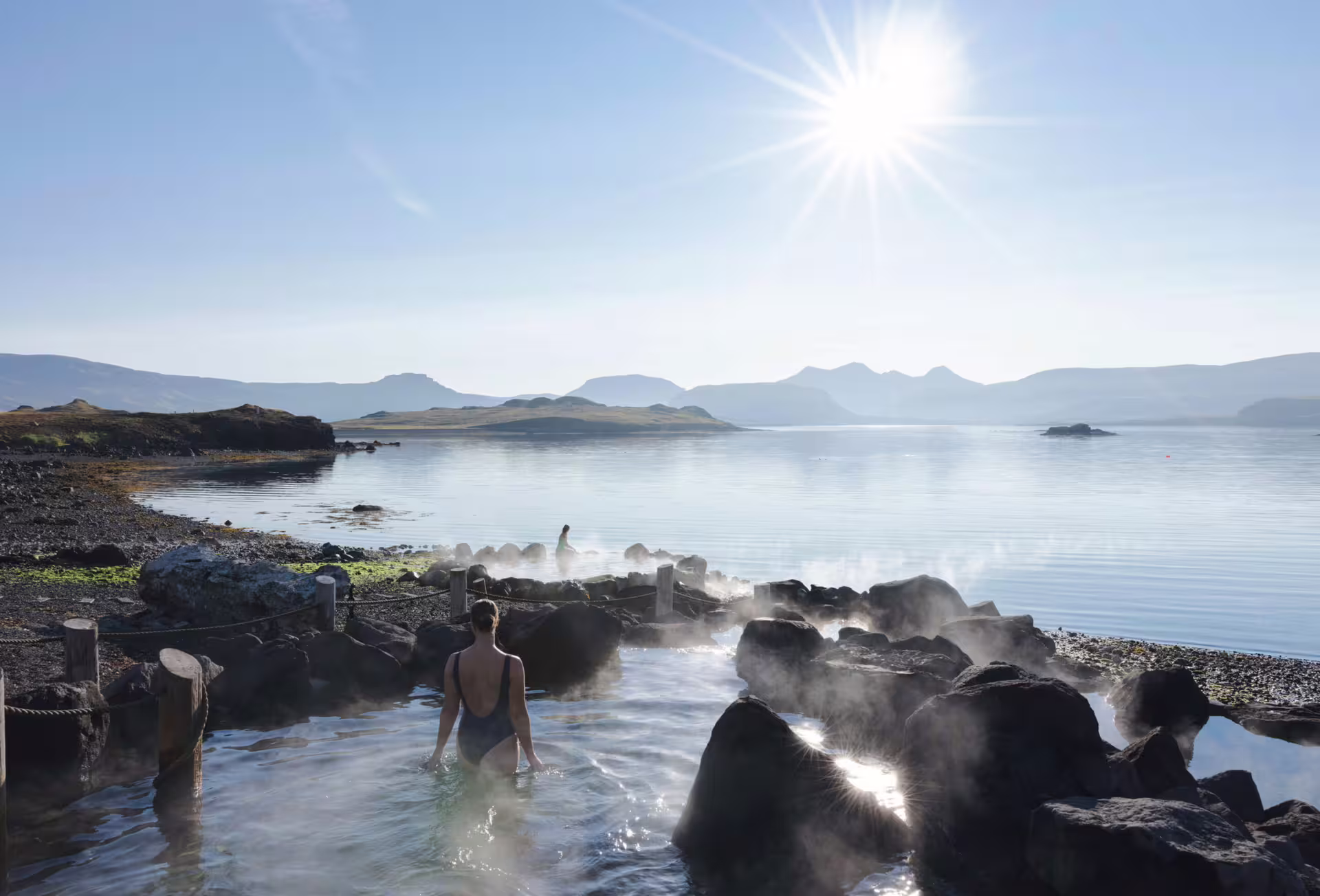 Woman soaking in a steaming geothermal hot spring beside a tranquil Iceland fjord with misty mountains and bright sunshine.