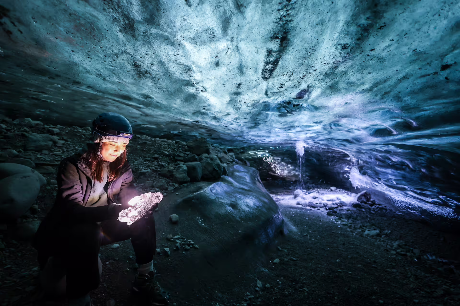 Woman with headlamp holds glowing ice crystal inside a blue Vatnajokull Glacier ice cave guided tour