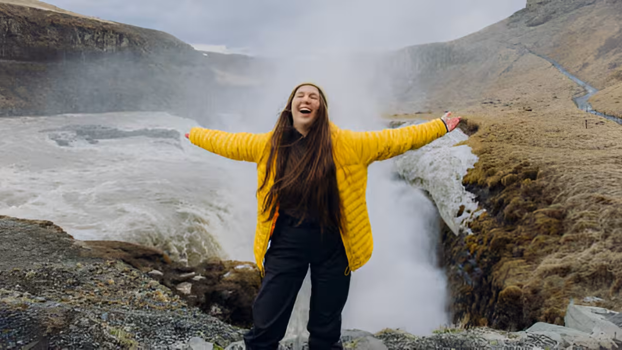 Happy woman in yellow jacket arms outstretched at Gullfoss waterfall on Iceland Golden Circle tour