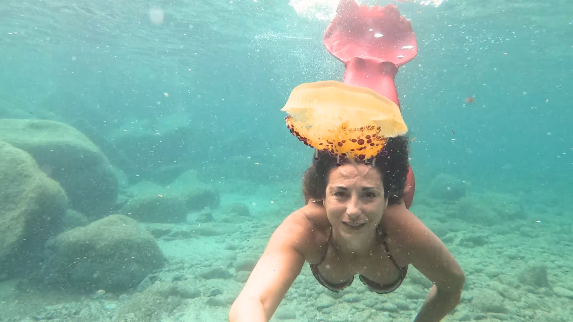 Underwater view of a woman in a mermaid tail swimming near a jellyfish in Cagliari's Gulf of Angels.