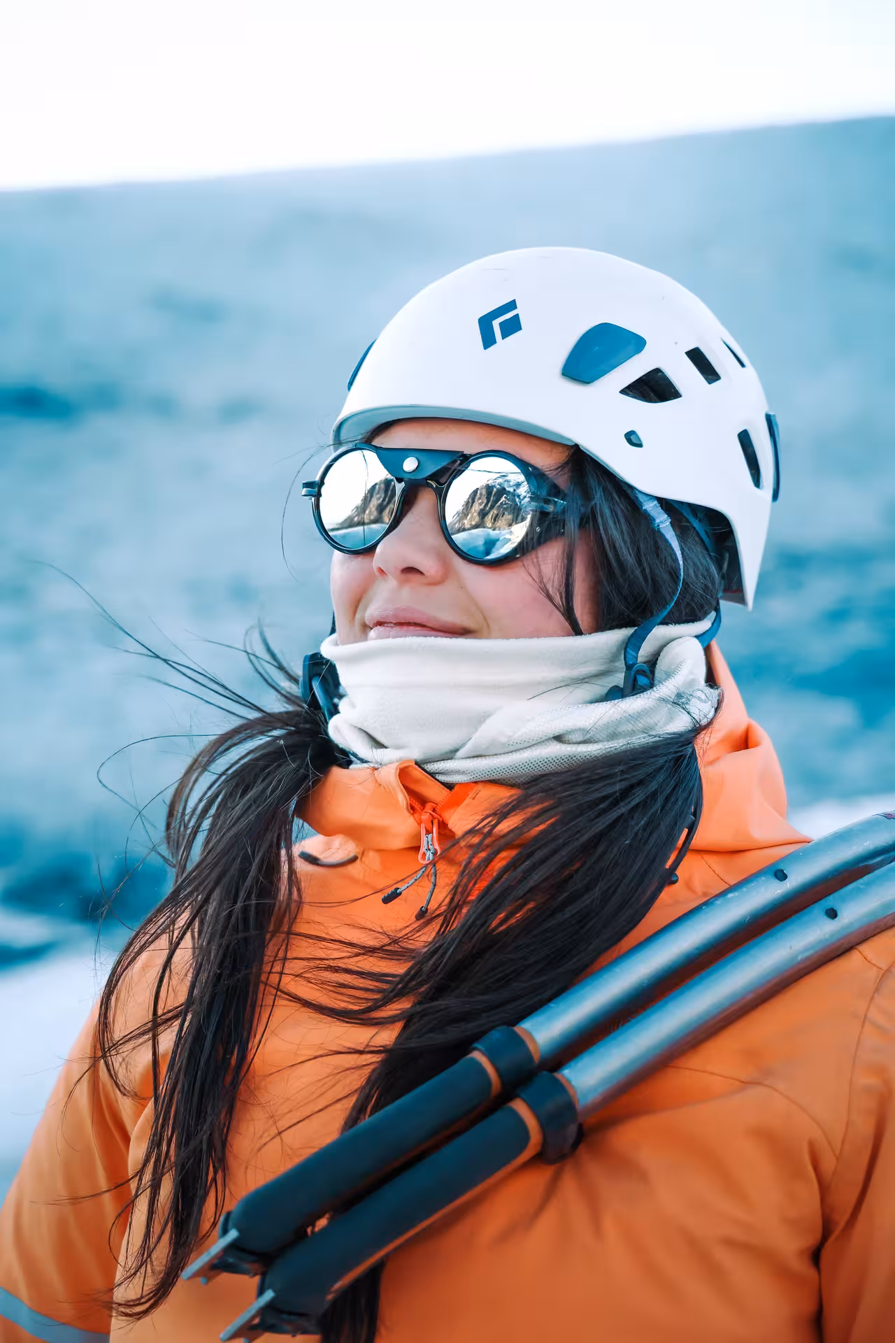 A smiling woman in an orange jacket and white helmet with glacier sunglasses ready for an Iceland winter zip line adventure tour.