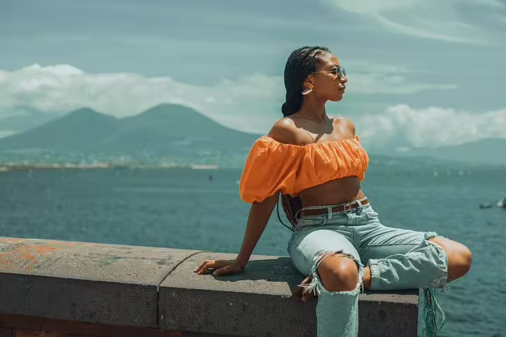 Woman in vibrant orange top sits on a seaside wall with Mount Vesuvius in the background during a Naples photoshoot.