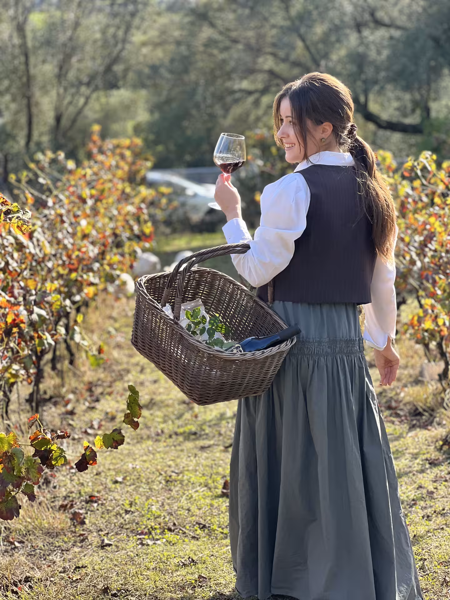 Woman with a basket and wine glass in hand, smiling in the scenic Olbia vineyard.