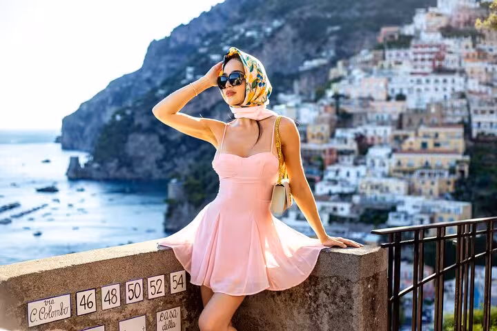 Stylish woman in a pink dress posing with Positano's colorful cliffs and azure sea, epitomizing Amalfi Coast elegance.