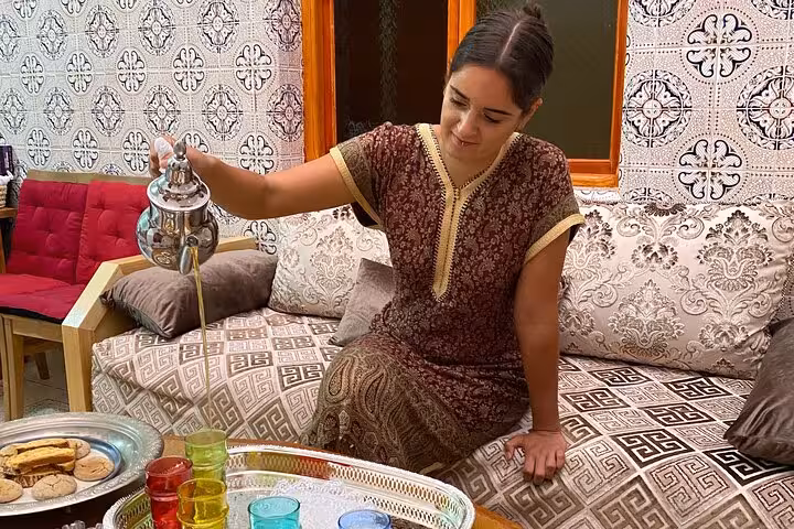 Woman in traditional attire pouring tea into colorful glasses during a cultural cooking class at a family home.