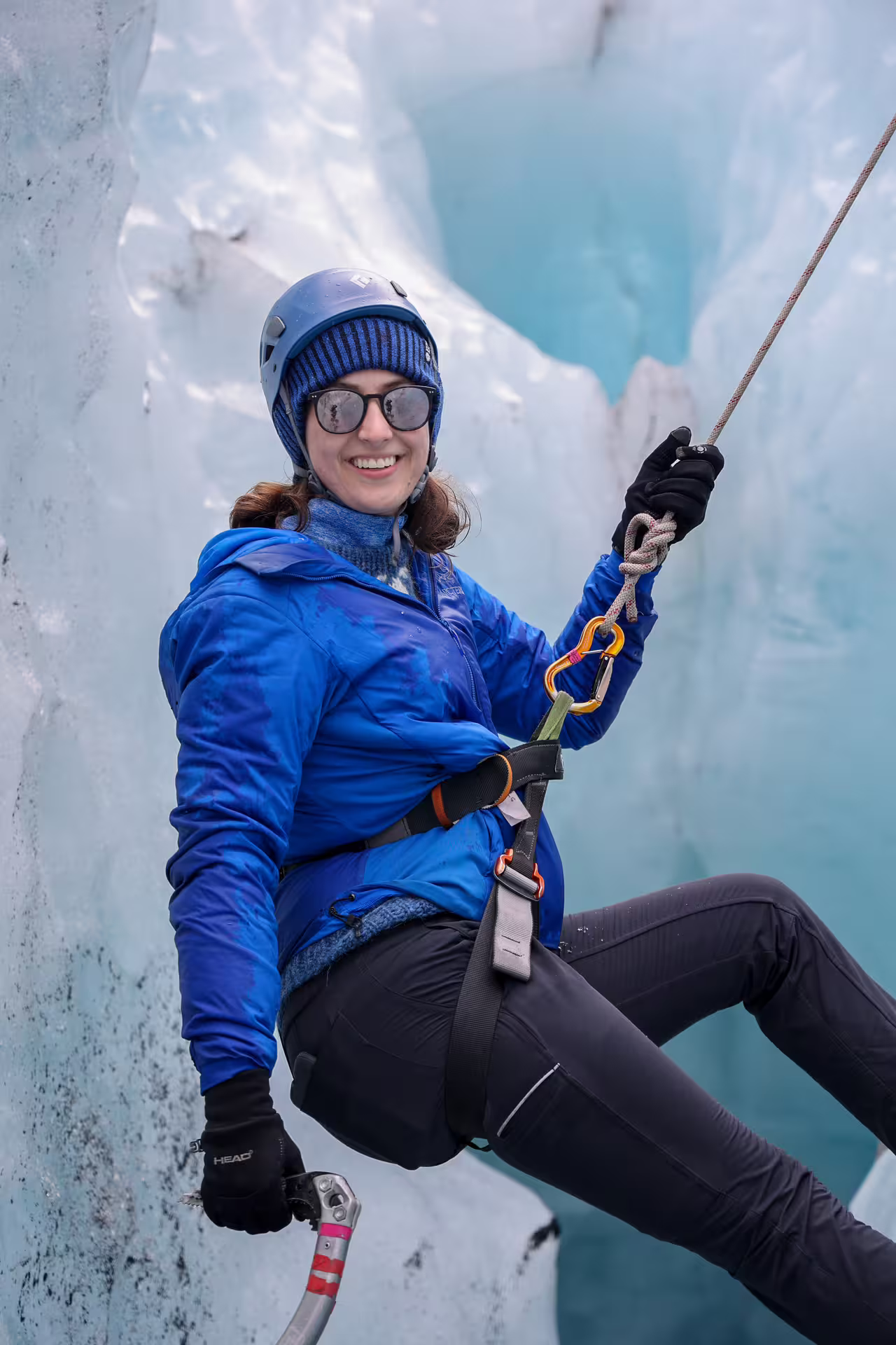 A smiling woman in blue jacket and helmet rappels down icy blue glacier wall on a private Solheimajokull ice climbing tour