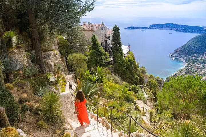 Woman in red dress descends stairs surrounded by lush greenery with panoramic views of Eze and the sea.