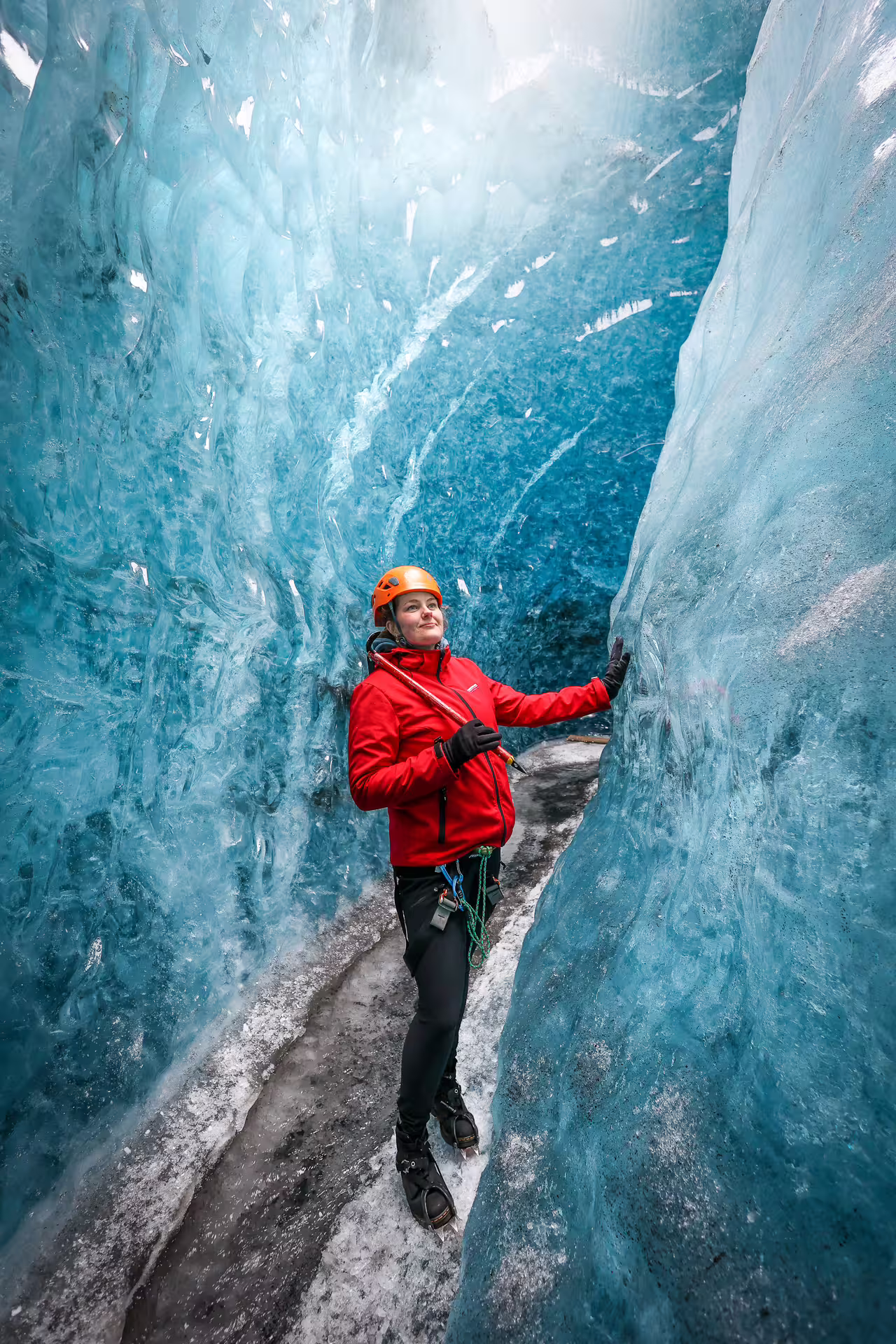 Woman in red jacket and orange helmet standing inside stunning blue ice canyon on Eggtrip extended glacier hike