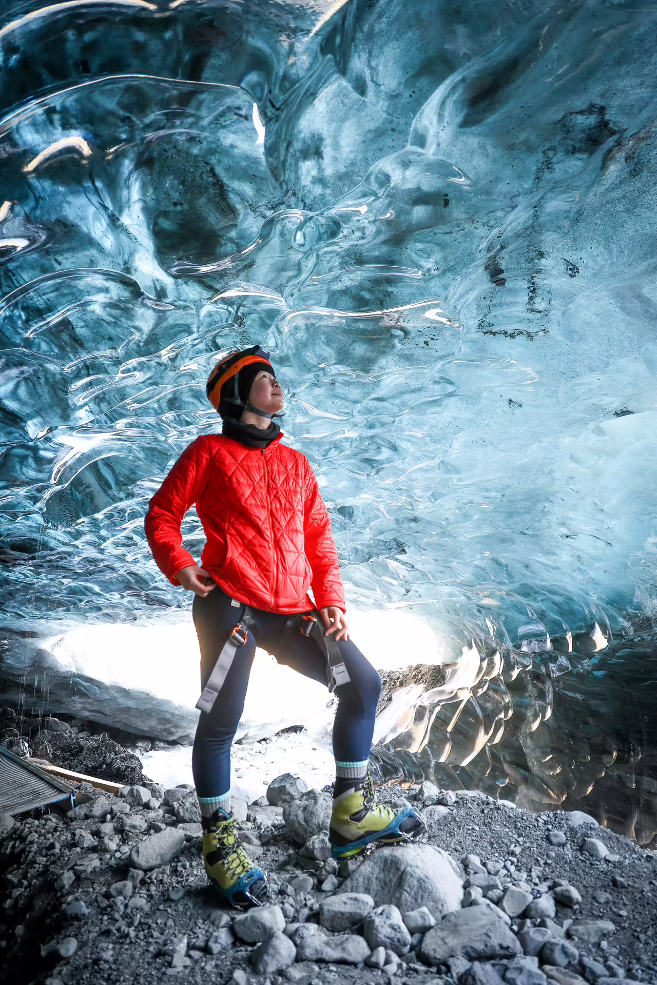 A woman in a red jacket and helmet marvels at the stunning blue ice ceiling inside an Icelandic glacier ice cave.