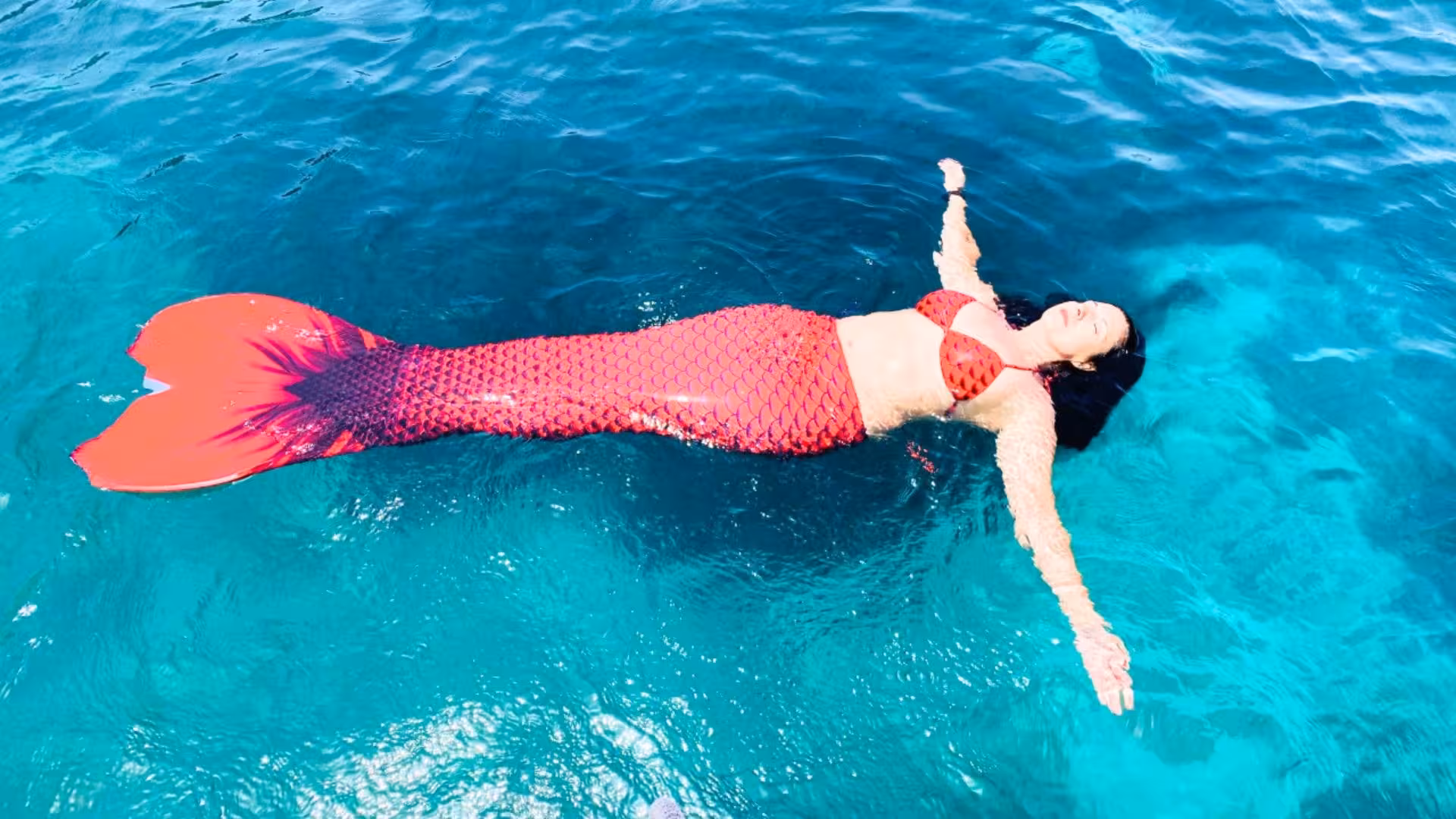 Woman in a red mermaid costume floating on clear blue waters in the Gulf of Angels, Cagliari experience.