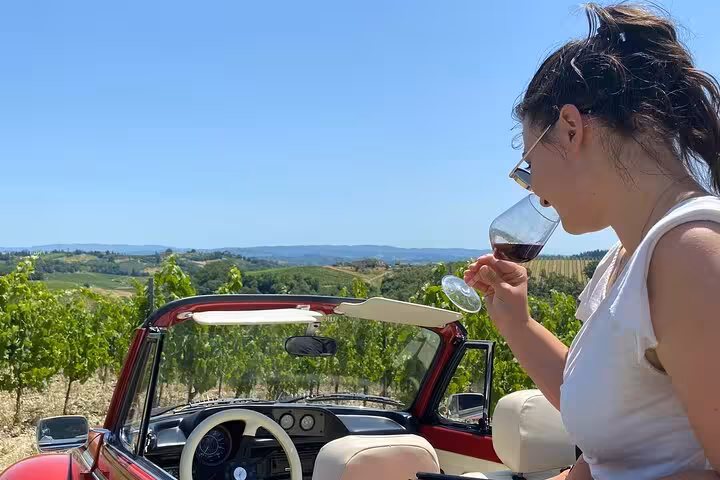 Woman savoring red wine by a vintage Beetle in Tuscany's scenic vineyard landscape.
