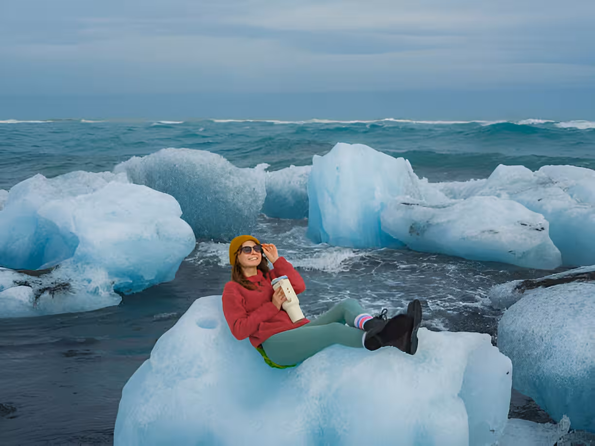 Woman sitting on glacier ice chunk at Diamond Beach Iceland with turquoise icebergs and ocean waves